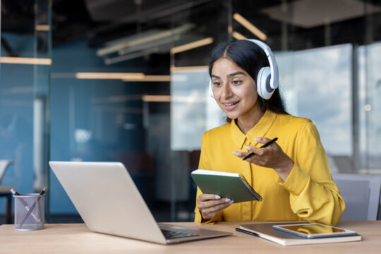 Smiling young Indian woman wearing headphones sitting at a desk in the office, talking on a video call on a laptop and taking notes in a notebook