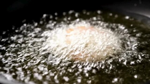 Close-up shot of food being deep-fried in sizzling hot oil, creating a golden, crispy texture. The cooking process shows vigorous bubbling as the item transforms into a delicious meal