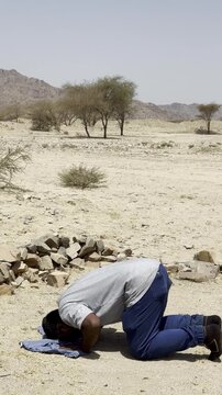 A Muslim man performs sujood prayer in a vast desert under a bright sky surrounded by rugged mountains and sparse vegetation symbolizing faith humility spirituality and devotion to Allah.