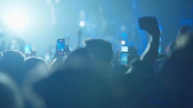 Silhouetted audience at a live music show films the performance on their smartphones. Footage transitions from bright, hazy lighting to a dark, atmospheric scene with blue stage lights.