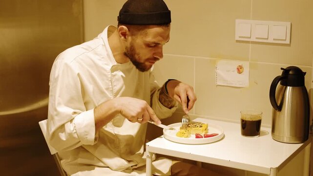 Professional baker eats omelet with tomatoes using fork and knife during a short break, sitting at a small table in an industrial bakery kitchen