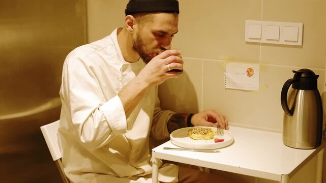 Professional baker drinks homemade compote during a short work break, sitting at a small table with omelet in an industrial bakery kitchen