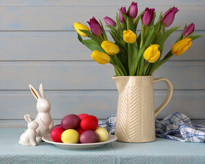 Festive Easter display with a variety of vibrant dyed eggs, cute ceramic bunny figures, and a vase overflowing with lively tulips on a soft blue wooden backdrop