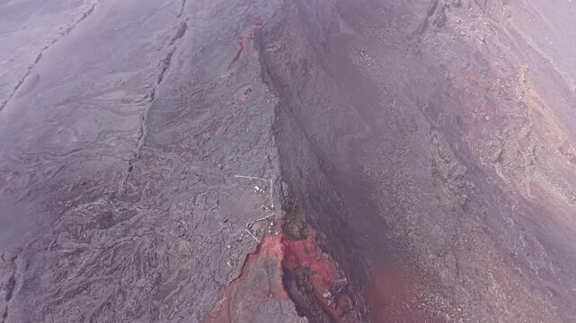 Drone footage flying along the rim of Dolomieu Crater at Piton de la Fournaise on Reunion Island. The shot reveals extensive volcanic rock formations, fractured lava layers and steep crater Walls.