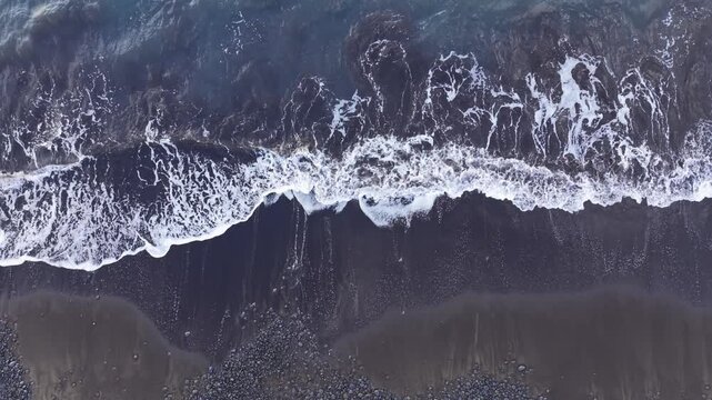 Top-down aerial drone footage showing a fixed overhead view of ocean waves breaking onto a dark shoreline. The shot emphasizes natural foam patterns, water movement.