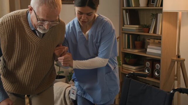 Young female nurse in medical scrubs holding senior man while helping him get into wheelchair in modern retirement home