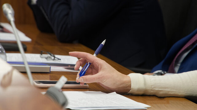 Female official sitting at wooden desk with colleagues during political summit. Hand with pen ready to sign contract or take notes near microphone system and eyeglasses. Photo
