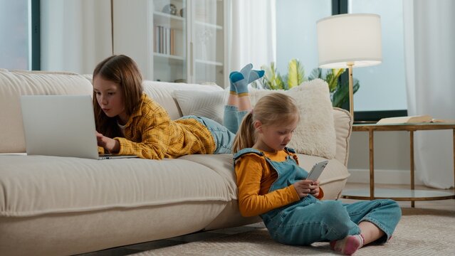 Two Caucasian little girls children using laptop and mobile phone gadgets addicted kids sisters siblings playing virtual video games browsing online internet technology devices at home living room