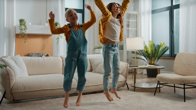 Two happy Caucasian sisters children kids singing song jumping dancing listening music in living room smile European siblings girls artistic daughters sing listen song dance jump together at home