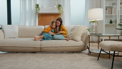 Naklejka premium Two Caucasian little kids girls sibling children reading book curious sisters read fairy tale watching pictures photos on couch sofa relaxing studying learning together study learn in living room home