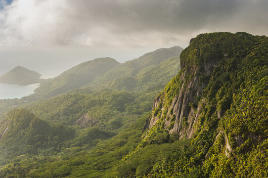 Aerial view of lush green mountains cascading down to the turquoise coast, framed by dramatic cloud formations, Seychelles, Seychelles.