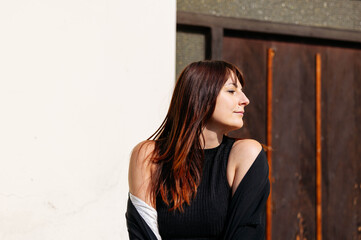 Fototapeta premium Young woman with long brown hair, wearing a black top and white jacket, poses confidently against a textured wall, showcasing a relaxed and stylish outdoor atmosphere
