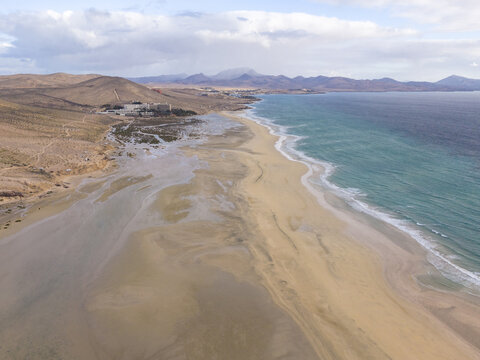 Aerial view of the turquoise sea meeting the sandy shores under a vast sky, where the coastline curves against the distant mountains, Jand&Atilde;&shy;a Natural Park, Fuerteventura, Canary Islands, Spain.