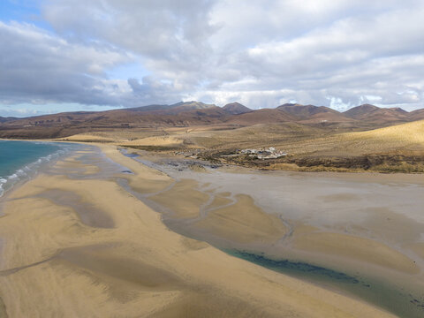 Aerial view of golden sands meeting turquoise waters under a vast sky, framed by rugged mountains in the distance, Jand&Atilde;&shy;a Natural Park, Fuerteventura, Canary Islands, Spain.
