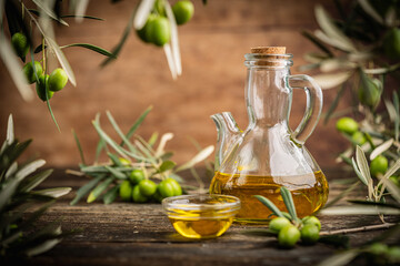 Obraz premium Glass bottle of olive oil with small bowl surrounded by fresh green olives and olive branches on wooden rustic table. Concept of Mediterranean cuisine, natural product, and organic cooking ingredient.