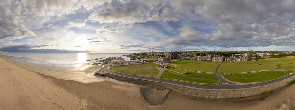 Aerial view of golden sands meeting the calm sea under a sky streaked with clouds, contrasting with the green links of St Andrews, Scotland, United Kingdom.