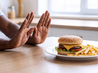 Person making stop gesture with hands to refuse cheeseburger and fries for healthy diet at home kitchen