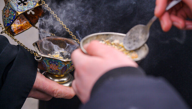 Priest's hand adding incense to a smoking censer during an Orthodox church ritual, rising aromatic smoke creating a solemn, sacred atmosphere of worship and prayer