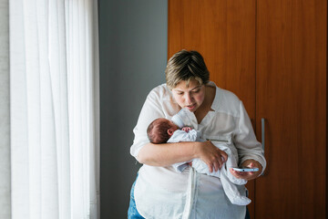 Mother holding newborn baby and using smartphone at home