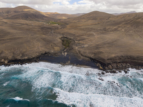 Aerial view of the rugged coastline where dark volcanic rocks meet the turquoise ocean, contrasting with the arid landscape, Playa de la Solapa, Fuerteventura, Spain.