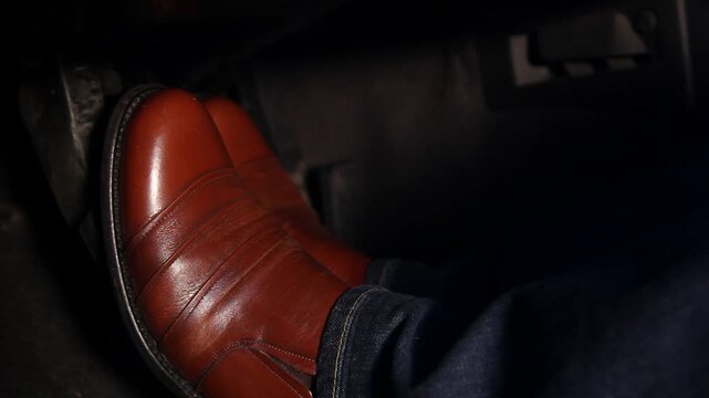 Close up view of a man's legs and feet wearing brown leather shoes and blue jeans while driving a car and pressing the brake and accelerator pedals.