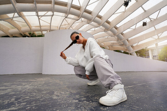 Hispanic woman wearing casual sportswear and sunglasses squatting under a white architectural structure