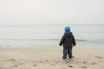 Child stands by the water looking at the ocean on a cloudy day at the beach with ships in the distance while waves touch the shore