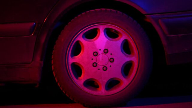 Close up of a car wheel illuminated by red and purple neon lights as it accelerates and drives away fast into the dark night city street scene.

