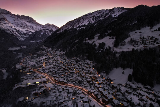 Aerial view of a snow-covered village nestled in a valley, illuminated by soft lights against a backdrop of majestic mountains, Champ&Atilde;&copy;ry, Valais, Switzerland.
