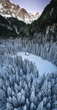 Aerial view of a solitary cabin nestled within a snow-dusted forest, framed by majestic, snow-capped mountains under a soft, pastel sky, Champ&Atilde;&copy;ry, Valais, Switzerland.