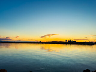 Obraz premium Sunset over calm water with silhouettes of boats and distant structures in a harbor scene at dusk