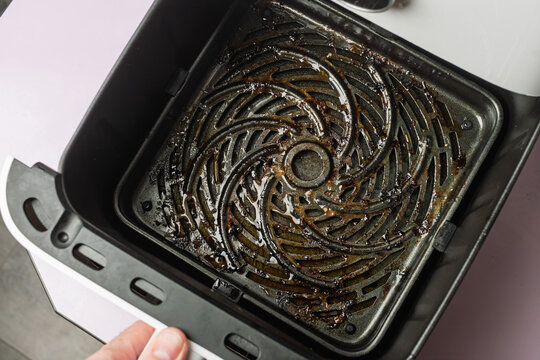 Hand pulls out dirty air fryer basket with burnt grease and food residue, concept of kitchen hygiene, appliance maintenance, cleanup.