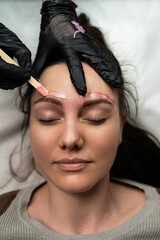 woman receiving eyebrow waxing treatment, gloved esthetician applying warm gel with wooden spatula over brow area, client eyes closed