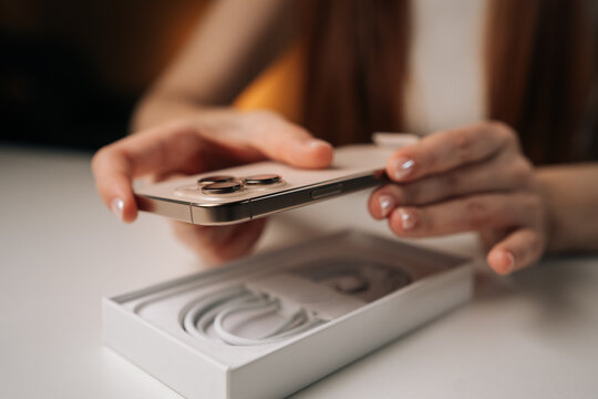 Female hands carefully removing new mobile phone from cardboard box, accompanied by charging cable, sitting at white table. Concept of anticipation and excitement of unboxing new device.