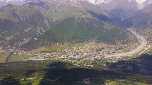Aerial drone pan and descent show Mestia or Ushguli on a braided river, patchwork fields, clustered houses, conifer forests, alpine meadows, and Mount Ushba in daylight