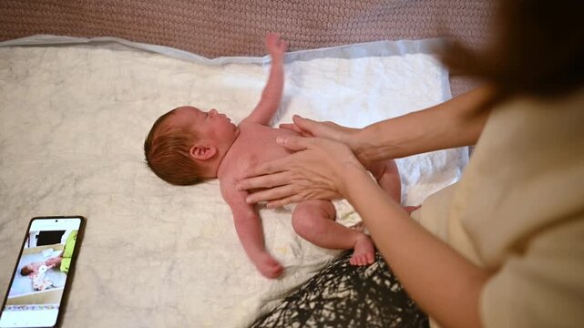 New mother gently massaging her infant's belly to relieve gas and aid digestion after a diaper change, while following instructions from a video tutorial on her smartphone nearby