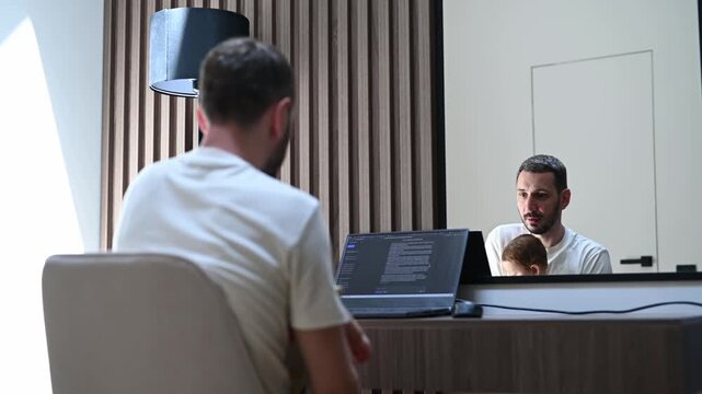 Young father and freelancer working on a laptop computer with his infant child sitting on his lap, trying to balance work and family life in a modern home office with a mirror view