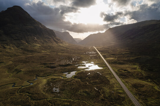 Aerial view of a ribbon of road cuts through the dramatic, shadowed valley of Glen Coe, under a sky pierced by radiant sunlight, Glen Coe, Scotland, United Kingdom.
