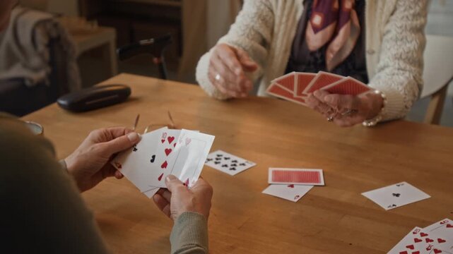 Over the shoulder shot of two unrecognizable women playing game of cards at wooden table in modern cozy retirement home