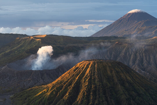 Aerial view of the rugged volcanic landscape, where the Bromo volcano rises amidst a sea of sand, its crater emitting a plume of smoke under a vast sky, Probolinggo, East Java, Indonesia.