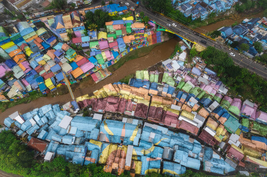Aerial view of a vibrant rainbow of houses cascading down to the river's edge, contrasting with the yellow bridge above, Malang, East Java, Indonesia.