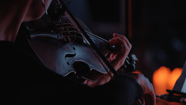 Intimate close-up of a violinist performing. Dramatic low-key lighting with warm, soft highlights illuminates the instrument hands. The moody, dark atmosphere suggests passion, focus, elegance