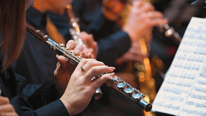 A focused close-up on a musician's hands playing a silver flute during a classical concert. Warm stage lighting highlights the instrument and player, with the orchestra blurred in the background