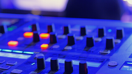 Close-up of a sound mixing board in atmospheric blue light. Glowing orange buttons contrast cool tones. Selective focus creates an abstract background for music, tech, or nightlife concepts
