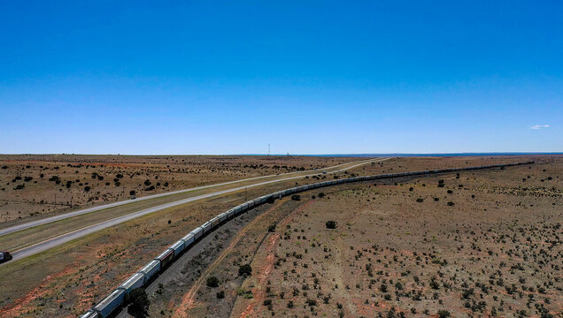 View of a seemingly endless freight train snakes through the arid landscape beside a long highway under a vast, clear blue sky, Winslow, Arizona, United States.