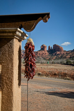 View of a vibrant chili ristra hanging under the eaves of a stucco building against the backdrop of the majestic red rock formations, Winslow, Arizona, United States.