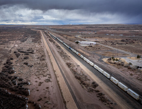 View of a long freight train stretching across the arid landscape under a stormy sky, a stark contrast of industry and nature, Winslow, Arizona, United States.