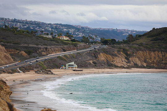 View of crashing waves meeting the sandy shore under a cloudy sky, a coastal road winding past bluffs topped with houses, Laguna Beach, California, United States.