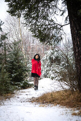 Asian woman wearing a red coat stands on a snowy path surrounded by evergreen trees in a winter forest setting with snow-covered ground and foliage