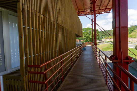 Perspective view of a modern wooden walkway with red steel railings, featuring a contemporary slatted timber facade and industrial architecture in a bright outdoor setting.